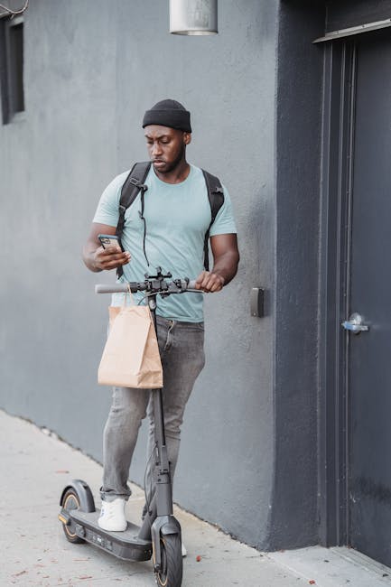 A young man with dark skin, wearing a black beanie, light blue T-shirt, grey jeans, and white sneakers, is standing on an electric scooter beside a grey building exterior. He is holding a brown paper 